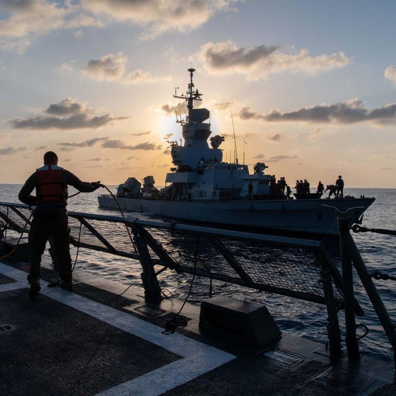 Naval personnel on the deck of a warship handle lines during a replenishment-at-sea or alongside operation with another naval vessel, silhouetted against a sunset over open ocean.
