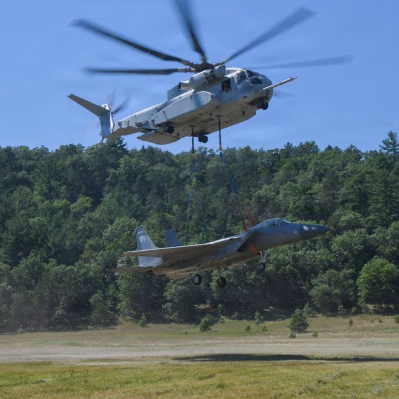 A CH-53K King Stallion heavy-lift helicopter carries an F-35 fighter jet airframe suspended by slings during an external lift operation over a grassy field with a tree line in the background.