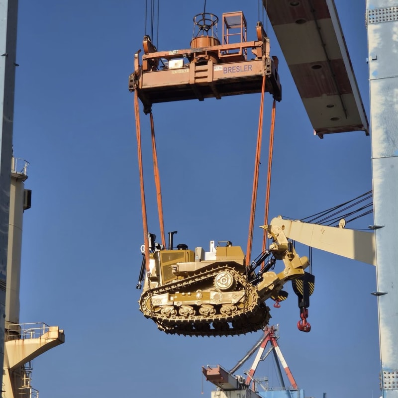 A yellow tracked bulldozer being lifted by a Bresler crane spreader at a port facility, suspended mid-air against a clear blue sky with port infrastructure visible in the background.