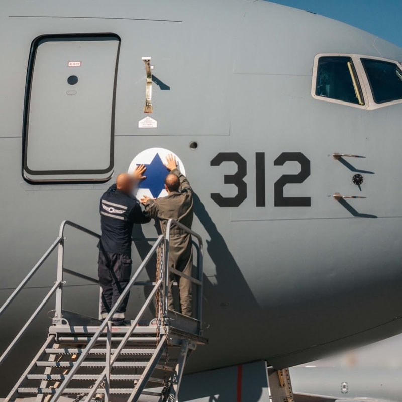 Two personnel on boarding stairs apply or inspect the Israeli Air Force roundel (Star of David insignia) on the nose of a military tanker/transport aircraft numbered 312, with another aircraft visible in the background.