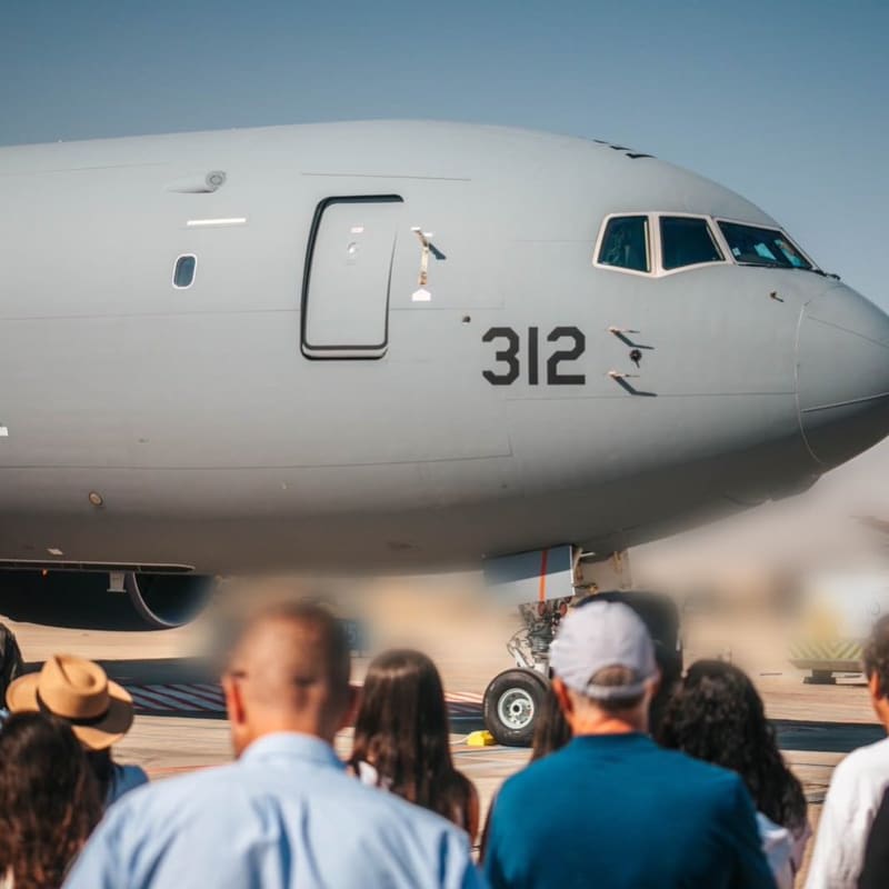 A crowd of spectators views the nose section of a gray military tanker/transport aircraft marked with the number 312, likely a Boeing KC-46 Pegasus, on a sunlit airfield tarmac during an open day or ceremony.