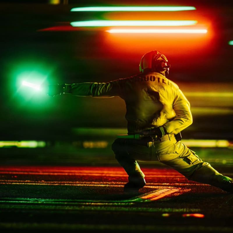 A catapult shooter on an aircraft carrier flight deck signals for launch during nighttime operations, crouching in the iconic launch pose with arm extended, illuminated by green and red deck lights with motion blur in the background.