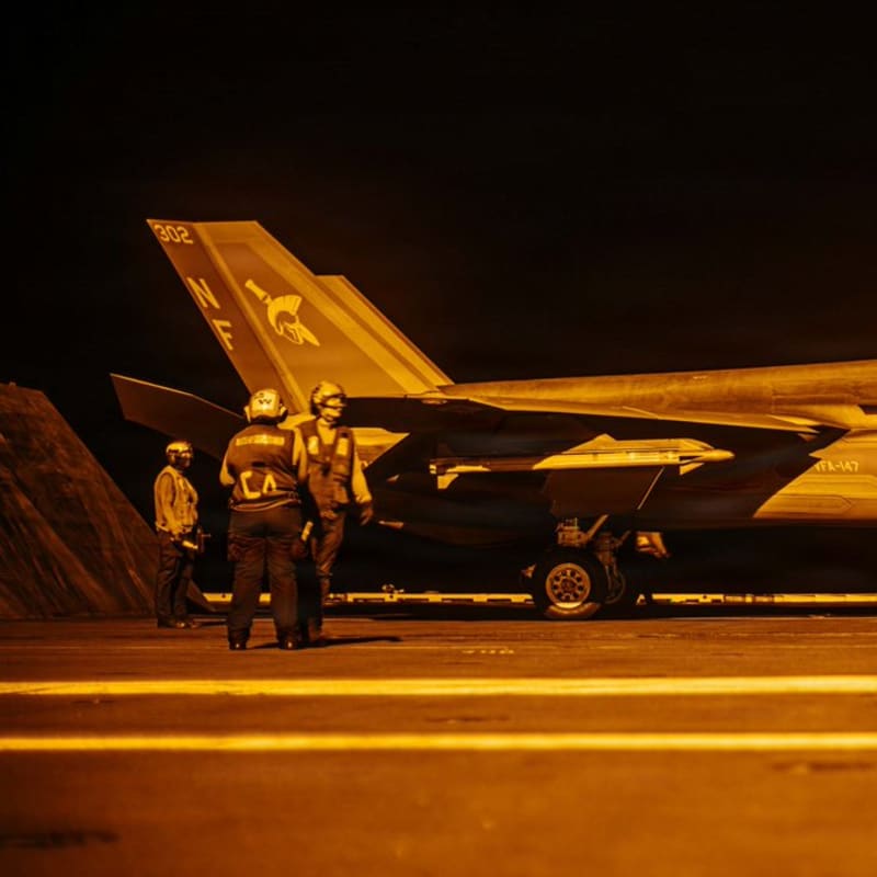 A U.S. Navy F-35C Lightning II from VFA-147 (tail code NF, number 302) sits on an aircraft carrier flight deck at night, bathed in amber lighting, with flight deck crew positioned around the aircraft.