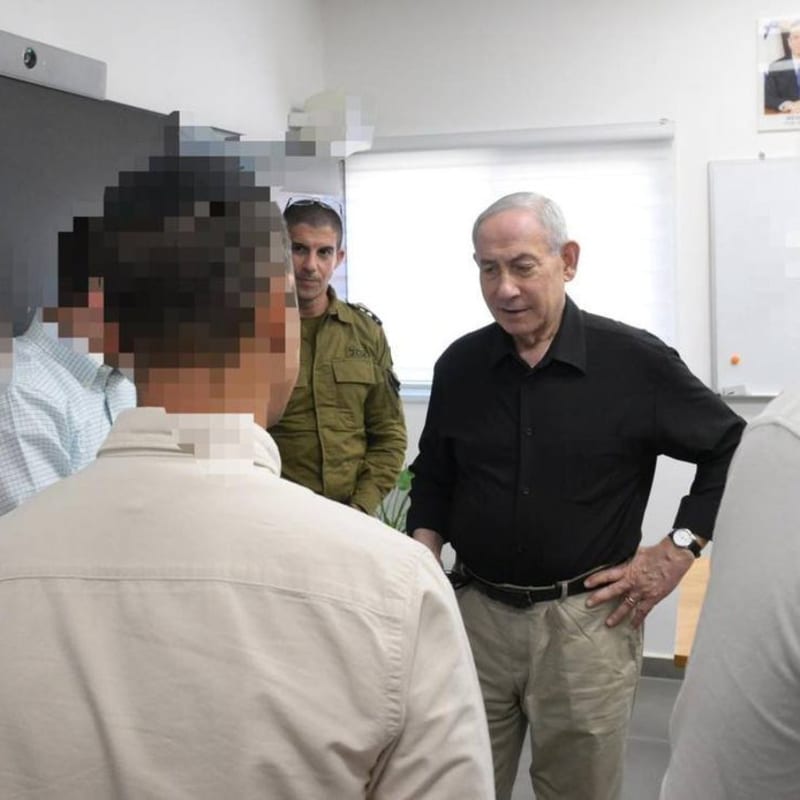 Israeli Prime Minister Benjamin Netanyahu, wearing a black shirt and khaki pants, meets with a group of individuals whose faces are pixelated for security reasons, accompanied by an IDF officer in uniform, in what appears to be a military or government briefing room.