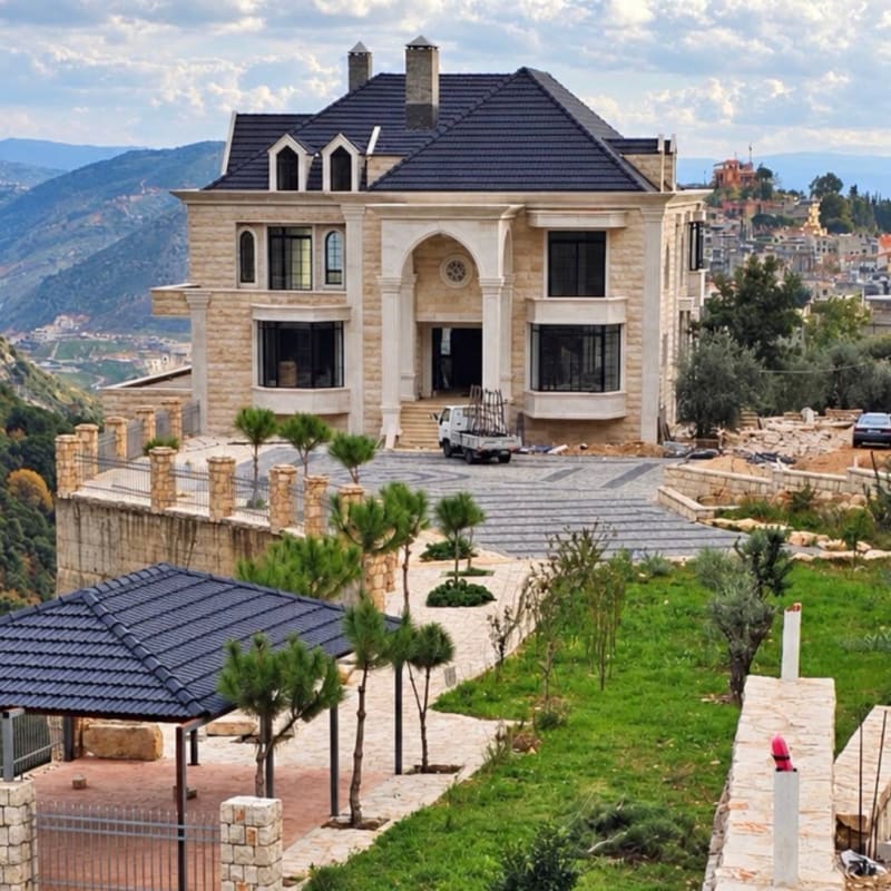 A large luxury stone villa with a dark blue tiled roof and grand arched entrance, situated on a hillside with landscaped gardens, likely in Lebanon based on the architectural style and mountainous terrain.