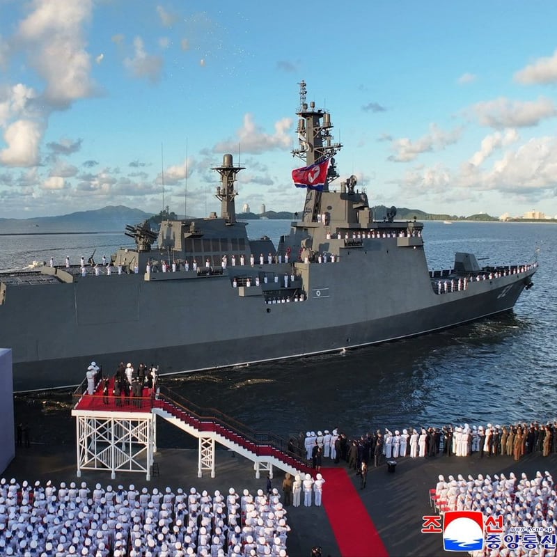 A North Korean naval warship flying the DPRK flag is displayed during a commissioning or launch ceremony, with rows of uniformed naval personnel standing at attention on the dock and aboard the vessel. A submarine is visible in the background, and the image bears the KCNA (Korean Central News Agency) watermark.