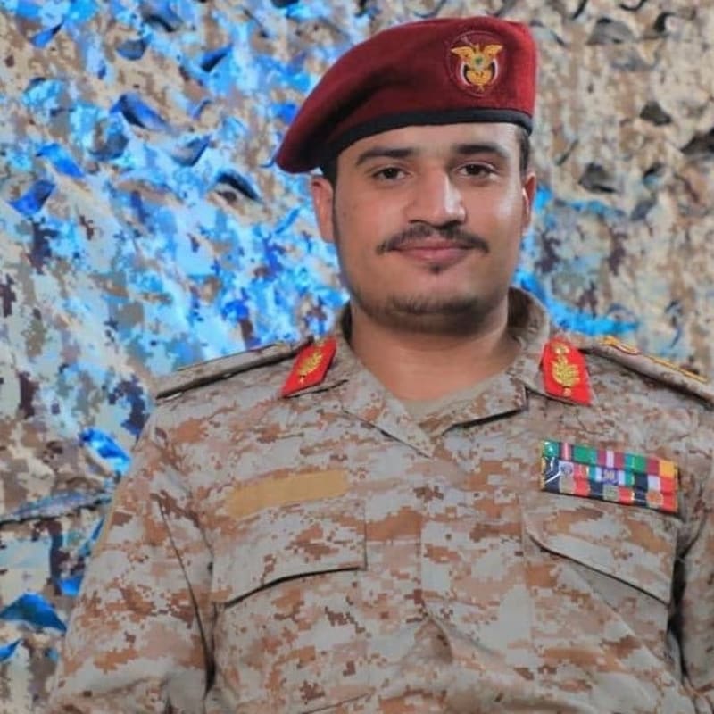 A military officer wearing a desert camouflage uniform with a maroon beret bearing an eagle emblem, military ribbons, and rank insignia, posed in front of a blue and tan camouflage netting backdrop, consistent with Yemeni military attire.