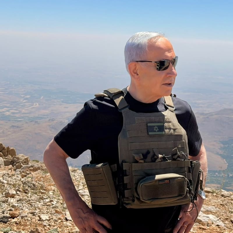 A man wearing sunglasses and a tactical vest with an Israeli flag patch stands on a rocky hilltop overlooking a vast arid landscape below.