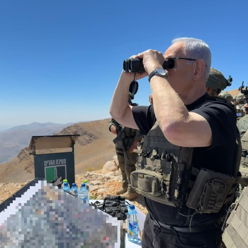 A man wearing a tactical vest looks through binoculars at a military observation point on a mountainous terrain, accompanied by soldiers. A sign with Hebrew text reading 'Mountain Brigade' is visible, and documents on the table are pixelated for security.