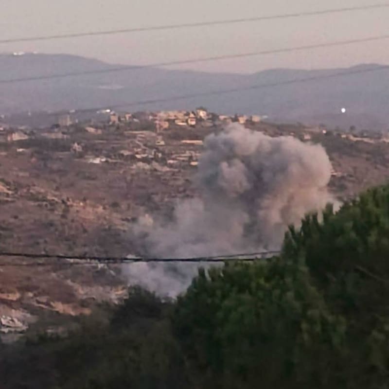 A large plume of smoke rises from a hillside in a rural, hilly landscape with scattered buildings visible in the background, consistent with an airstrike or explosion in southern Lebanon.