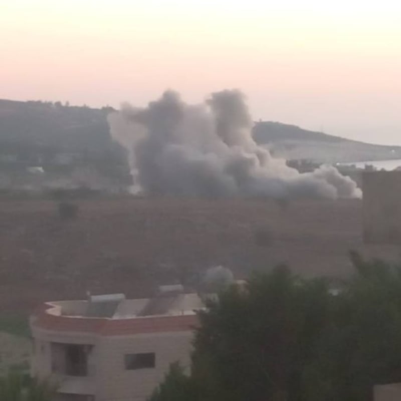 Smoke plumes rising from an airstrike or explosion in a hilly rural area with residential buildings in the foreground, likely in the Middle East.