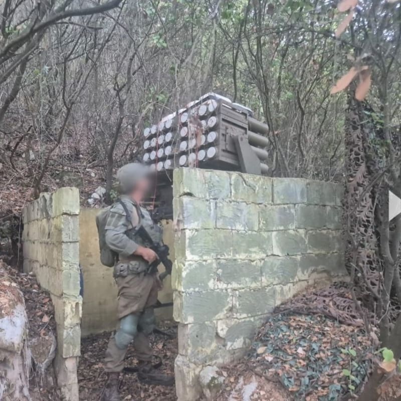 A soldier with a blurred face stands next to a concealed multiple rocket launcher system hidden behind a cinder block structure in dense woodland, likely depicting a Hezbollah rocket launcher position discovered in southern Lebanon.