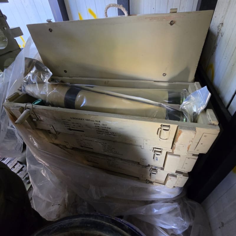 Stacked military ammunition crates labeled '2 ROCKET 107mm H.E.' dated 2017, with the top crate open revealing 107mm high-explosive rockets wrapped in protective packaging.