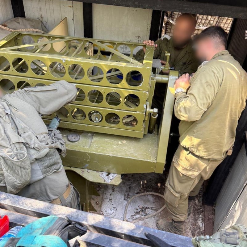 Soldiers with blurred faces in olive-drab uniforms inspect or handle a green multi-tube rocket launcher rack, likely a captured or seized multiple rocket launcher system, inside a confined space or vehicle.