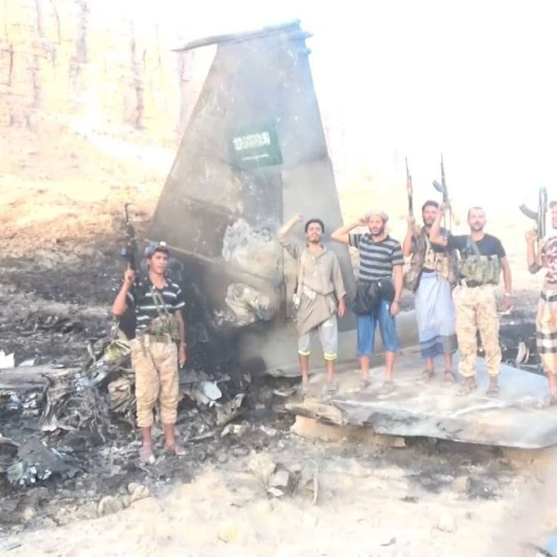 Armed fighters pose in front of the wreckage of a downed aircraft bearing Saudi markings in an arid landscape, with Arabic-language Yemeni military media watermark visible in the corner.