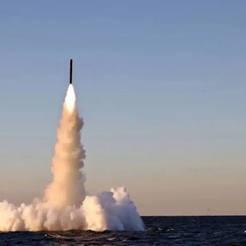 A submarine-launched ballistic missile emerging from the ocean surface with a large plume of smoke and exhaust trailing behind it against a clear sky.