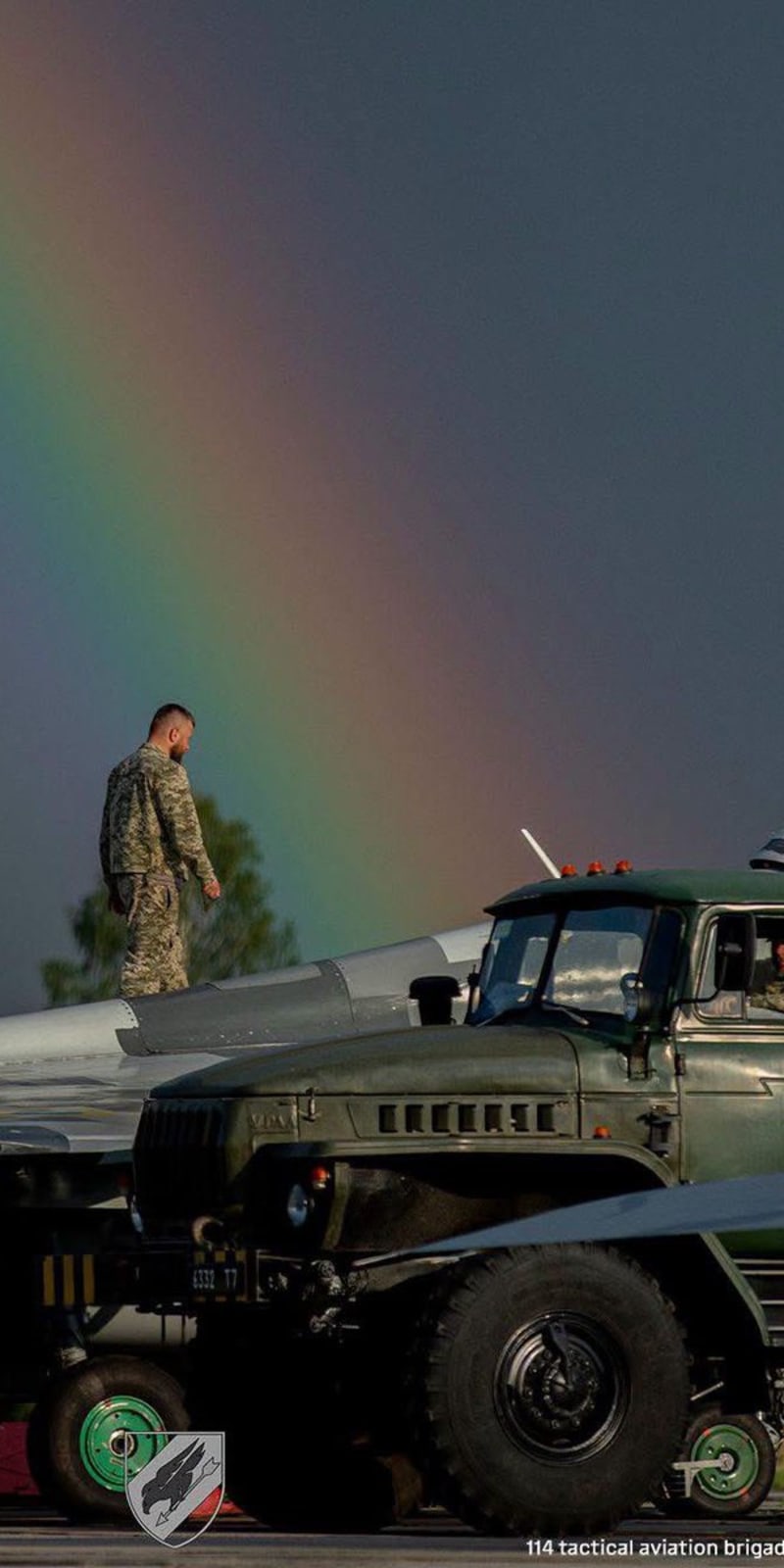 A Ukrainian military serviceman walks on the wing of a fighter jet on an airfield tarmac, with a military truck nearby and a vivid rainbow arcing across a dramatic sky. The image is credited to the 114th Tactical Aviation Brigade.