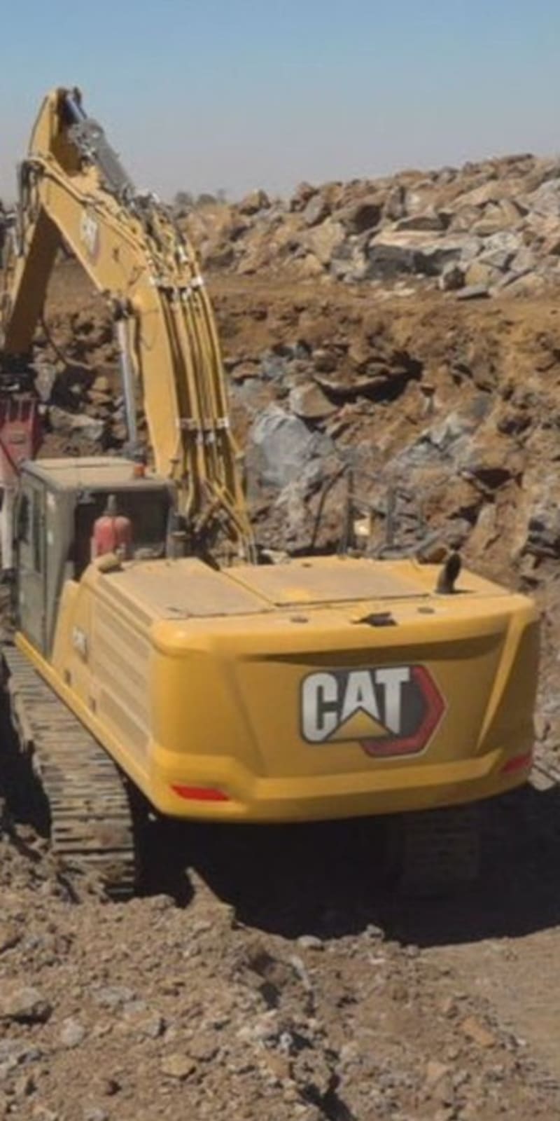 A CAT excavator digging a trench at a rocky construction or mining site in an arid landscape, with additional heavy machinery visible in the background.