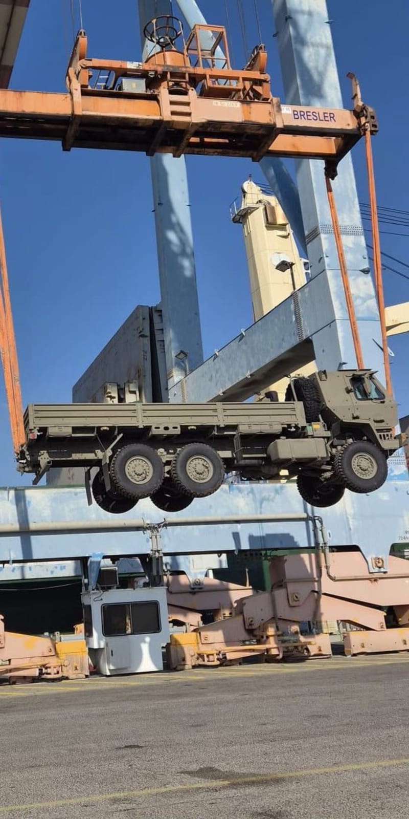 A port crane lifts a military truck at a commercial port, with a worker in a high-visibility vest and hard hat observing from below. The crane bears the name 'BRESLER'.
