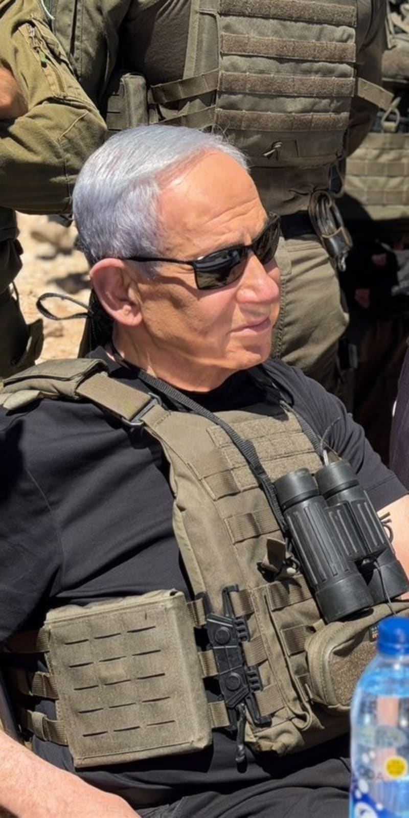 Two senior officials wearing tactical body armor and dark clothing sit surrounded by soldiers in military gear. One holds binoculars and wears sunglasses, suggesting a field visit or military briefing location.