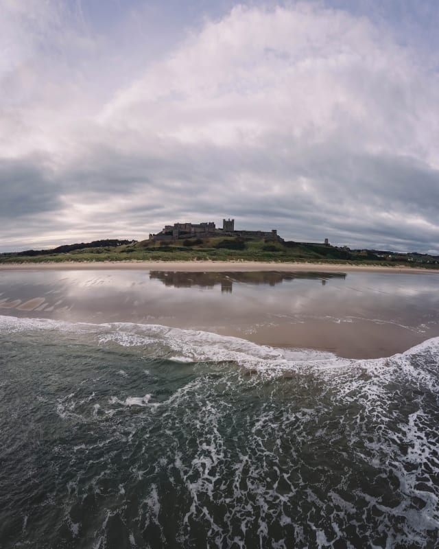 A drone photo of Bamburgh Castle. It is taken from out at sea. There are waves in the foreground and the castle is on the horizon reflecting perfectly in the wet sand on the beach.