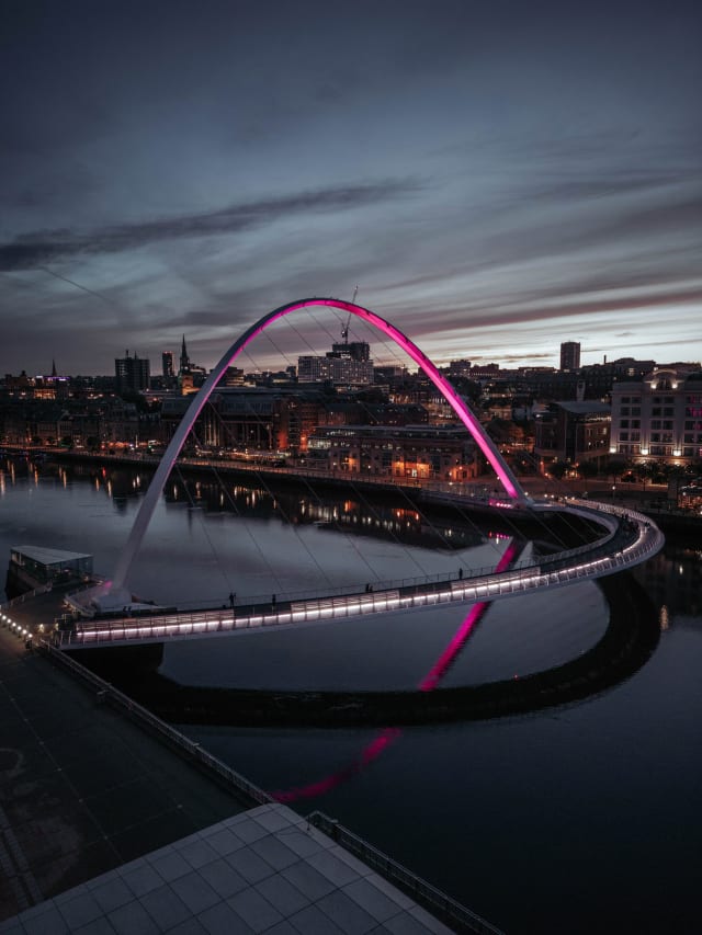 A drone photo of the Millennium Bridge in Newcastle. It is from the Gateshead side with the Newcastle Quayside in the background. The bridge is lit up with a pink light and the reflections in the water are mirror-like.