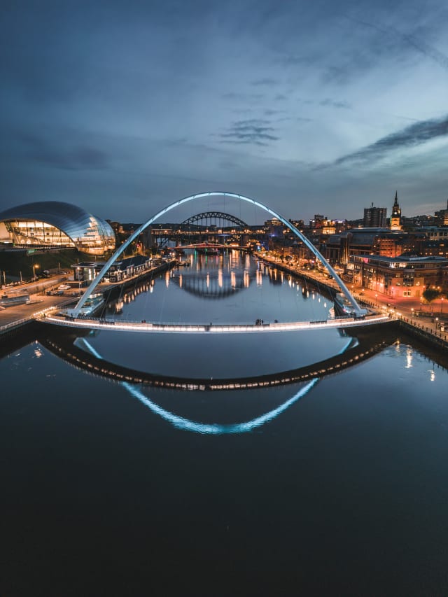 A drone photo of the Millennium Bridge in Newcastle. It is taken over the river tyne looking up-river. The arch of the Tyne Bridge lines up with the arch of the Millennium bridge, and the water is producing mirror-like reflections.