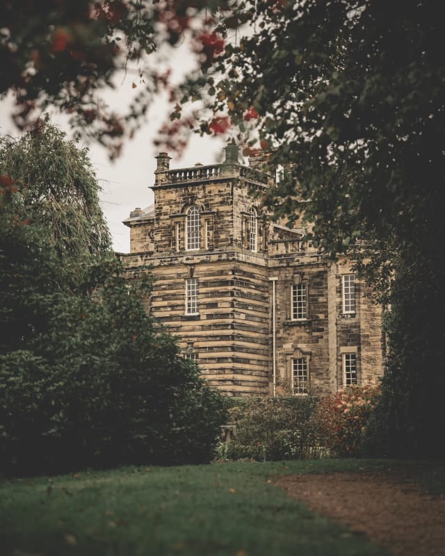 A photo through trees of Deleval Hall. It shows the west side of the building, there are trees on the left and right of the photo and a small gravel path running towards it.