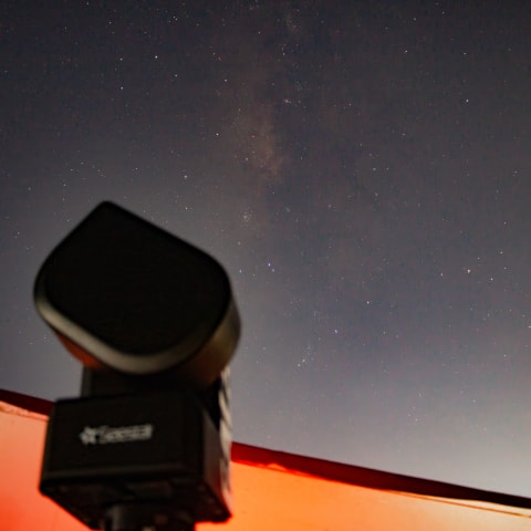 A faint band of the Milky Way rising through a starry sky, with a camera tracking mount in silhouette against an orange-lit roof edge