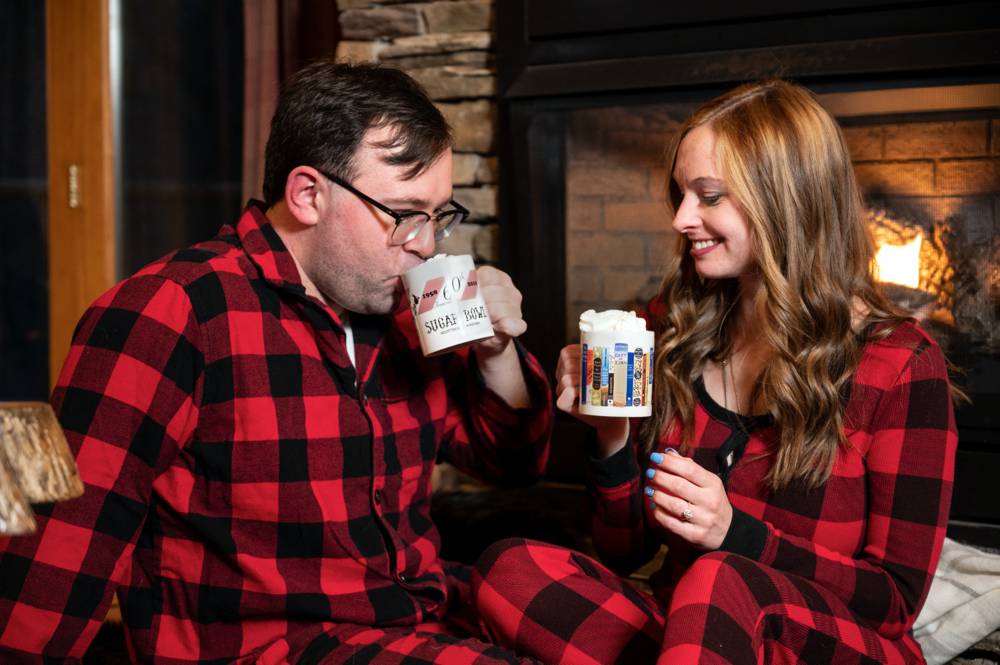 couple in front of fireplace in cabin drinking hot chocolate