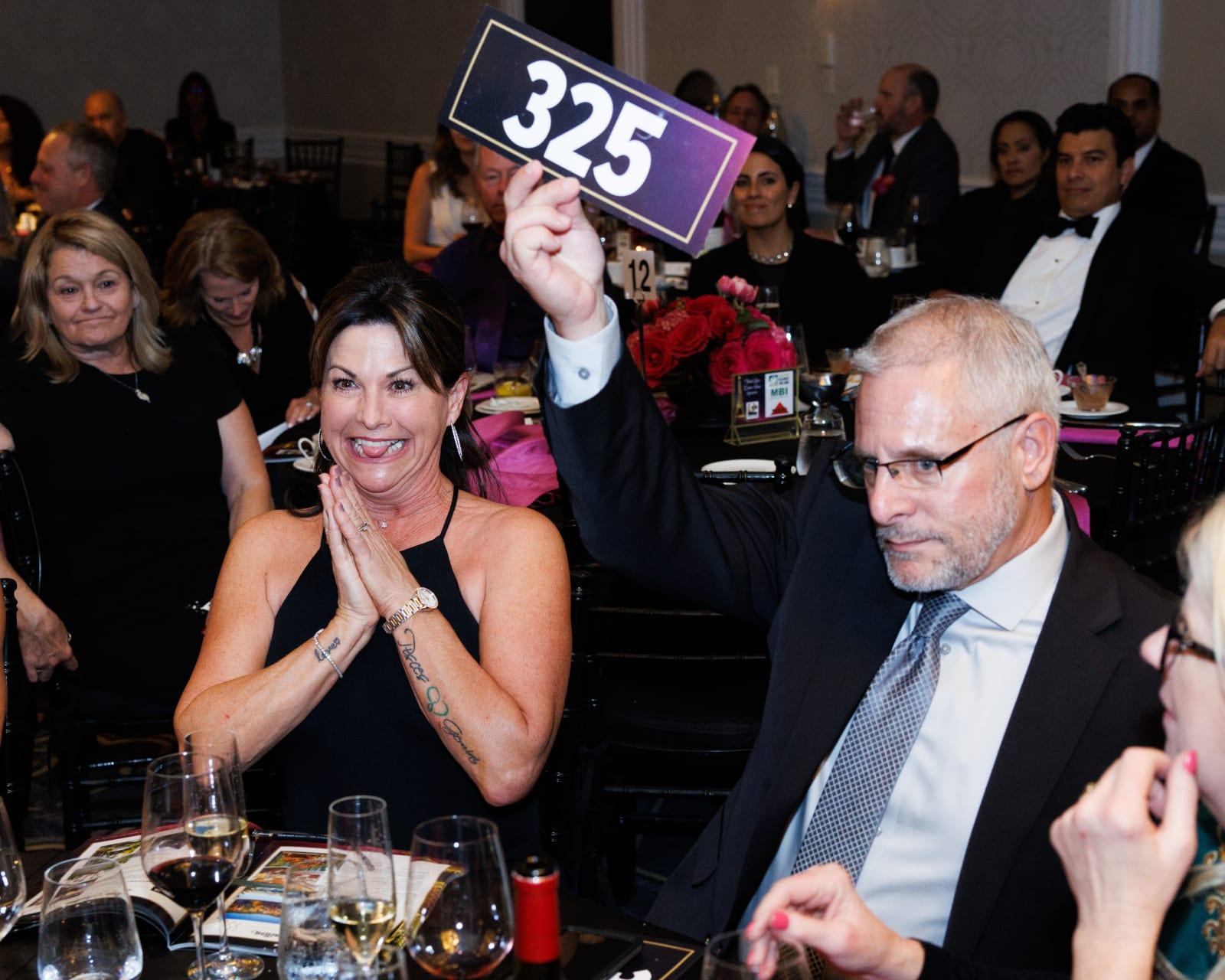 A woman claps excitedly beside a bidder raising paddle #325 high above the table, framed by pink and red floral centerpieces, during live bidding at the 2023 UCON Advocacy Auction.