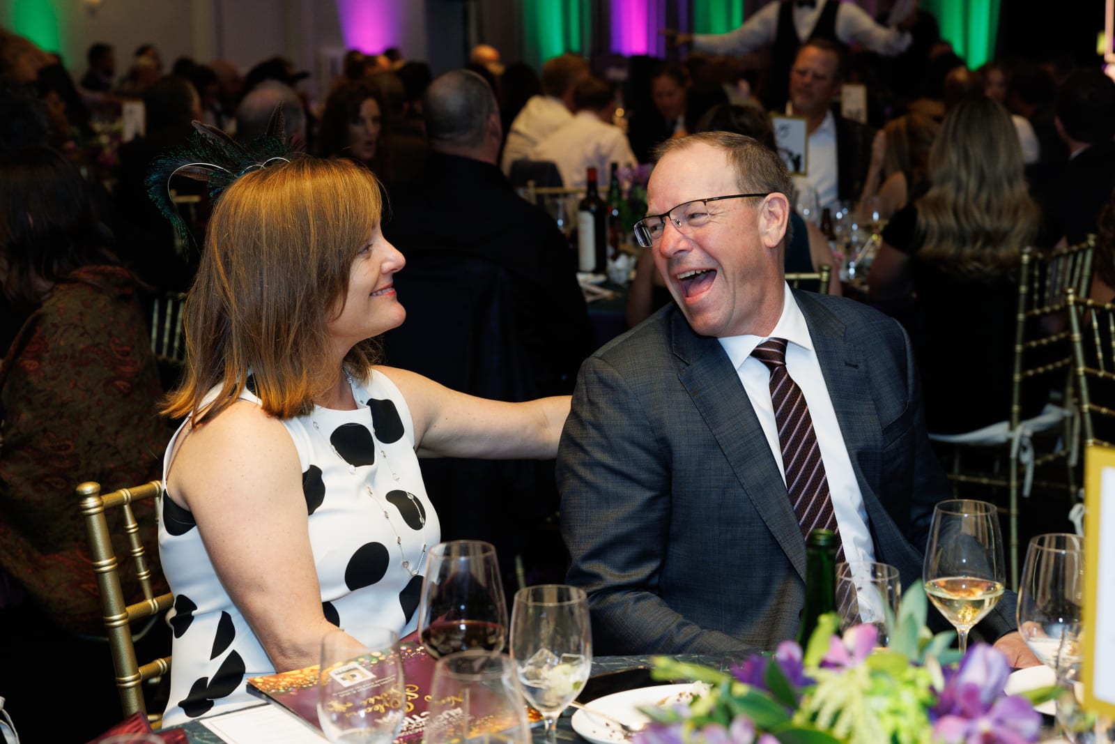 A woman rests her hand on a fellow guest's shoulder as they share a warm laugh at the dinner table, colorful green and purple lighting behind them, 2025 Advocacy Auction