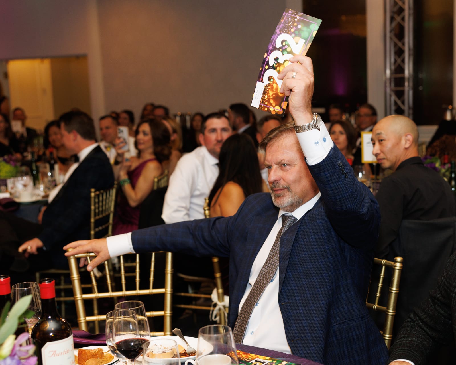 A guest in a navy suit dramatically raises his bid paddle overhead in an exciting moment during live bidding at the 2025 UCON Advocacy Auction.