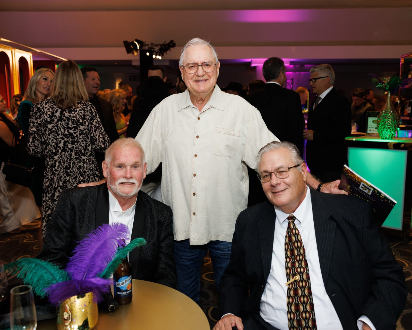 Three distinguished guests pose together at the 2025 UCON Advocacy Auction, with a purple feather centerpiece and colorful ambient lighting behind them.