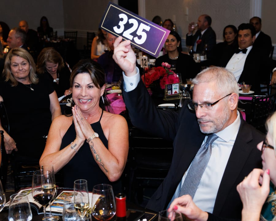 A woman claps excitedly beside a bidder raising paddle #325 high above the table, framed by pink and red floral centerpieces, during live bidding at the 2023 UCON Advocacy Auction.