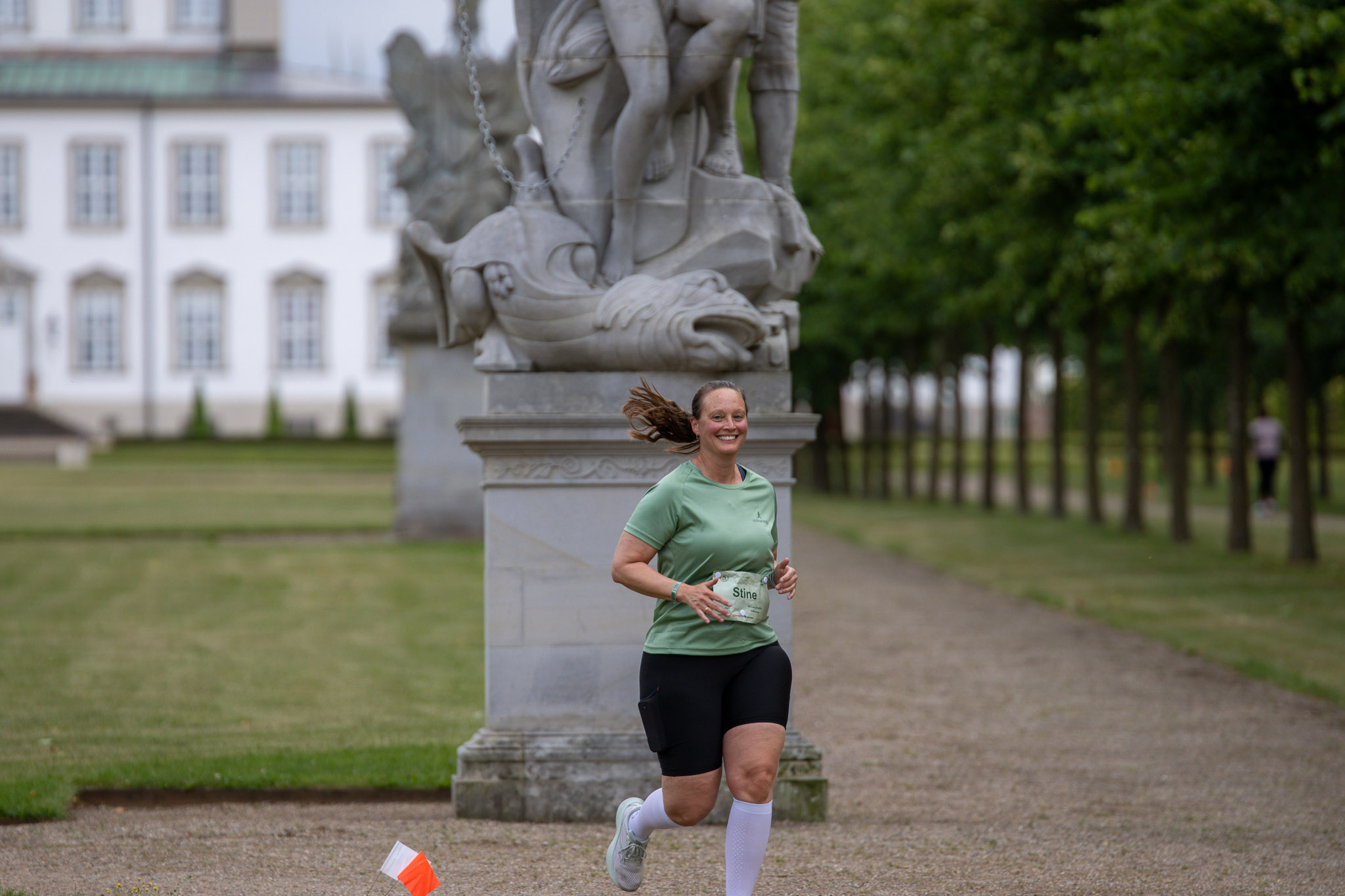En glad kvindelig løber foran en flot skulptur i Fredensborg slotspark med slottet i baggrunden.