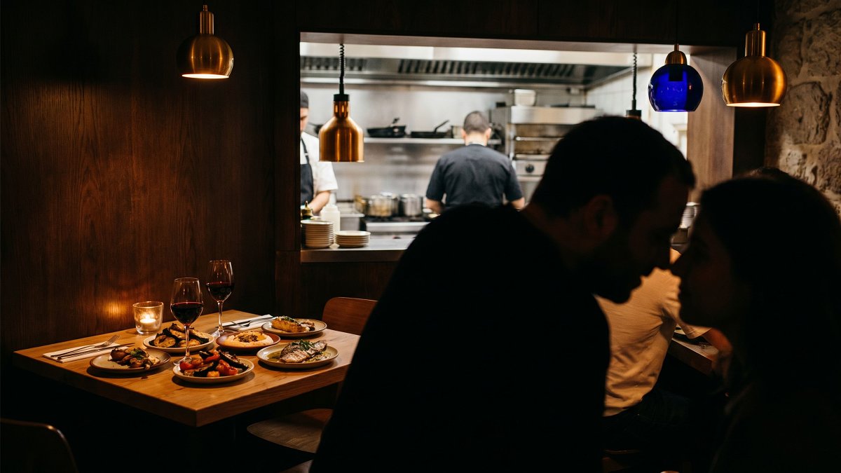 An intimate corner two-top in a low-lit kosher bistro in Tel Aviv, tea light glowing, two wine glasses of Galilee red, small plates of Mediterranean mezze, warm wood and brass interior