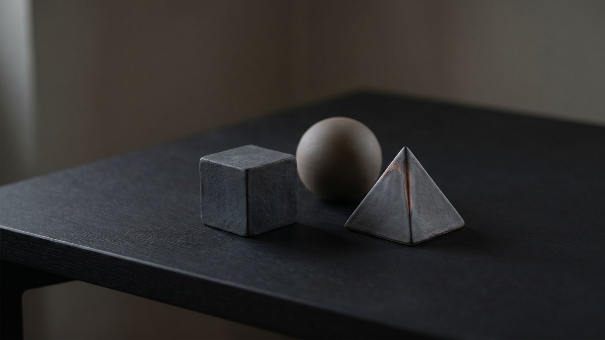 Three matte ceramic objects cube sphere pyramid arranged on a minimalist black desk casting long shadows