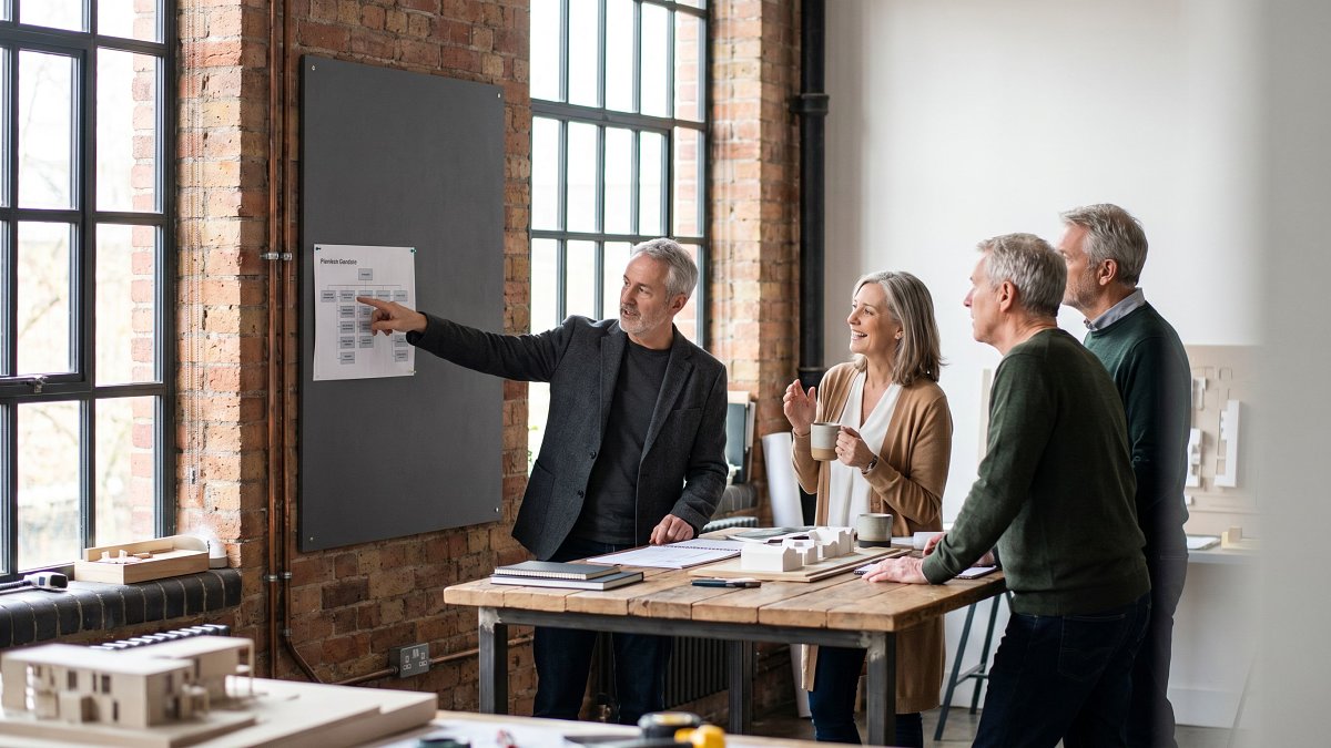 Small senior engineering team of four around a high standing desk in a renovated atelier, mid-morning light through steel windows