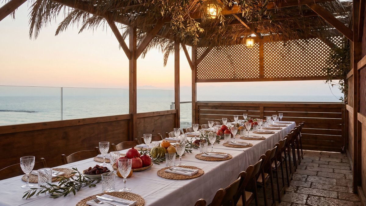 An oceanfront kosher Sukkot sukkah on a Tel Aviv hotel terrace at dusk, set for chol hamoed lunch