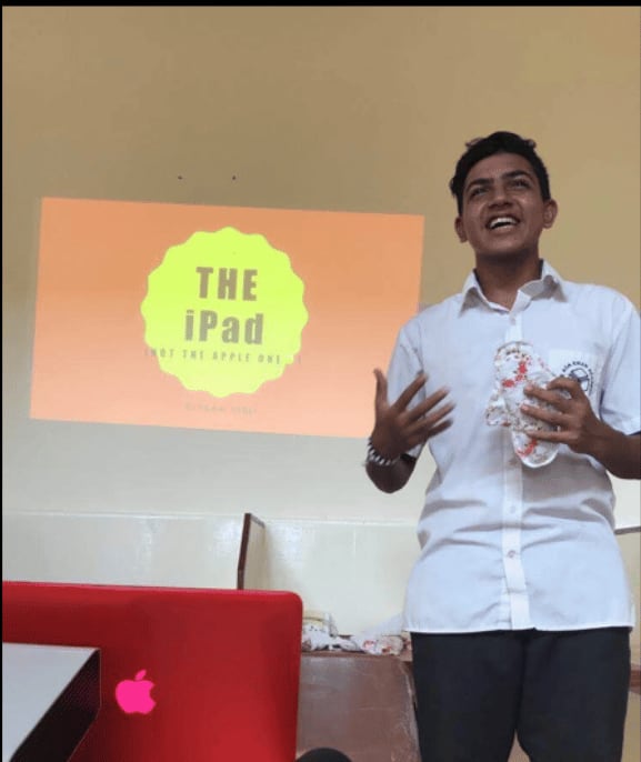 A teenage Ziyaan Virji presenting to a classroom, holding a reusable menstrual pad, in front of a projected slide reading THE iPad, not the Apple one