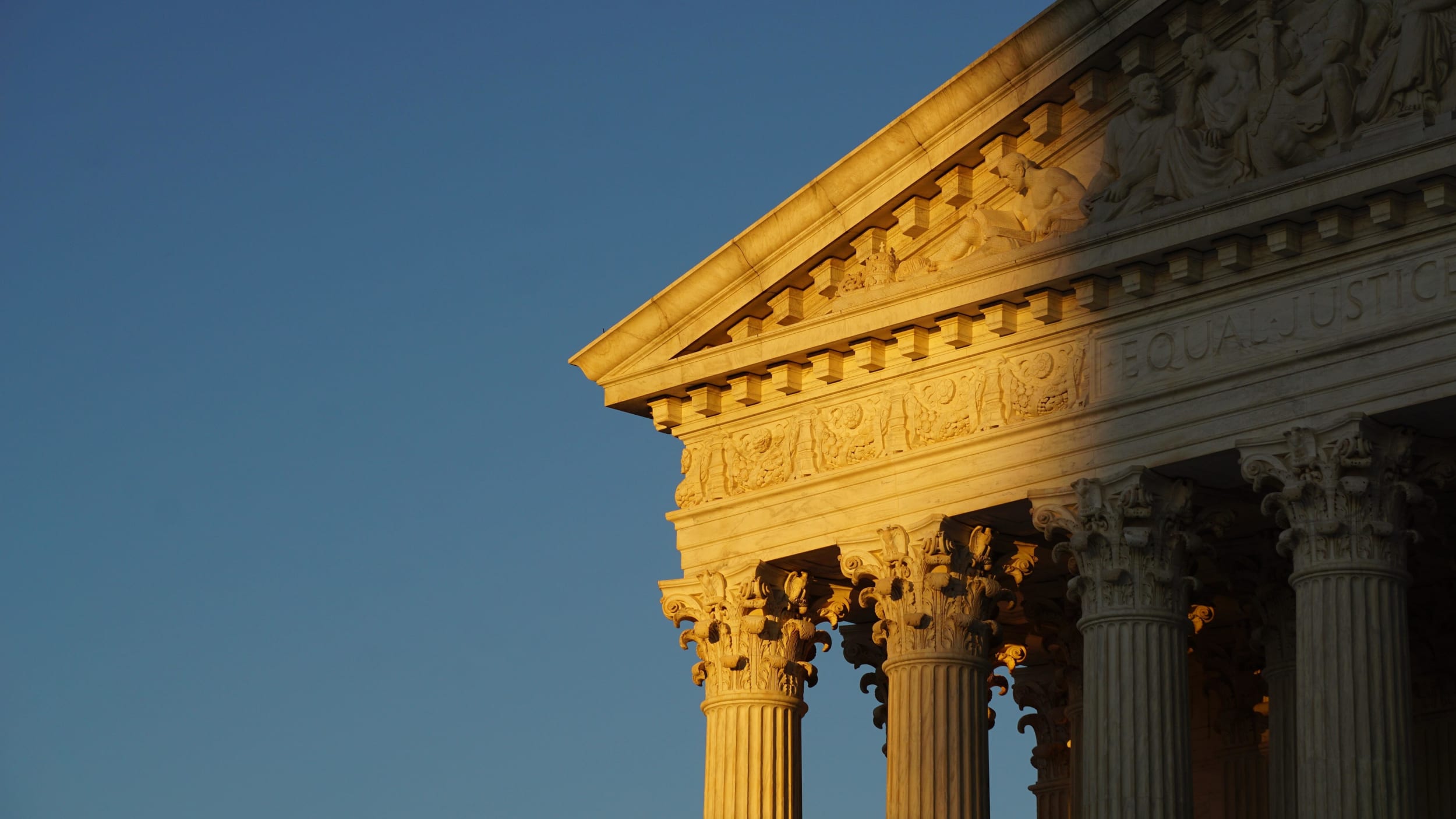 Photo of the top of the U.S. Supreme Court building Washington D.C., half covered in shadow and half in golden light with a blue sky background.