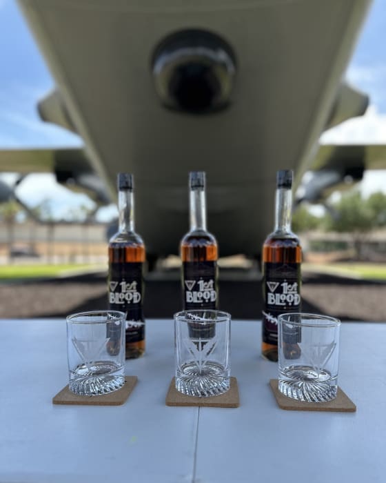 Three bottles of 1st Blood and three glasses set out beneath a gunship.