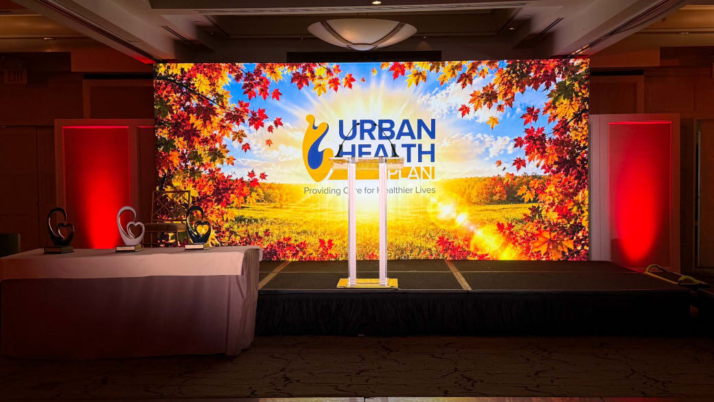Backlit Urban Health Plan stage backdrop of autumn leaves over a sunlit field, with a clear acrylic podium on stage and heart-shaped awards on a side table