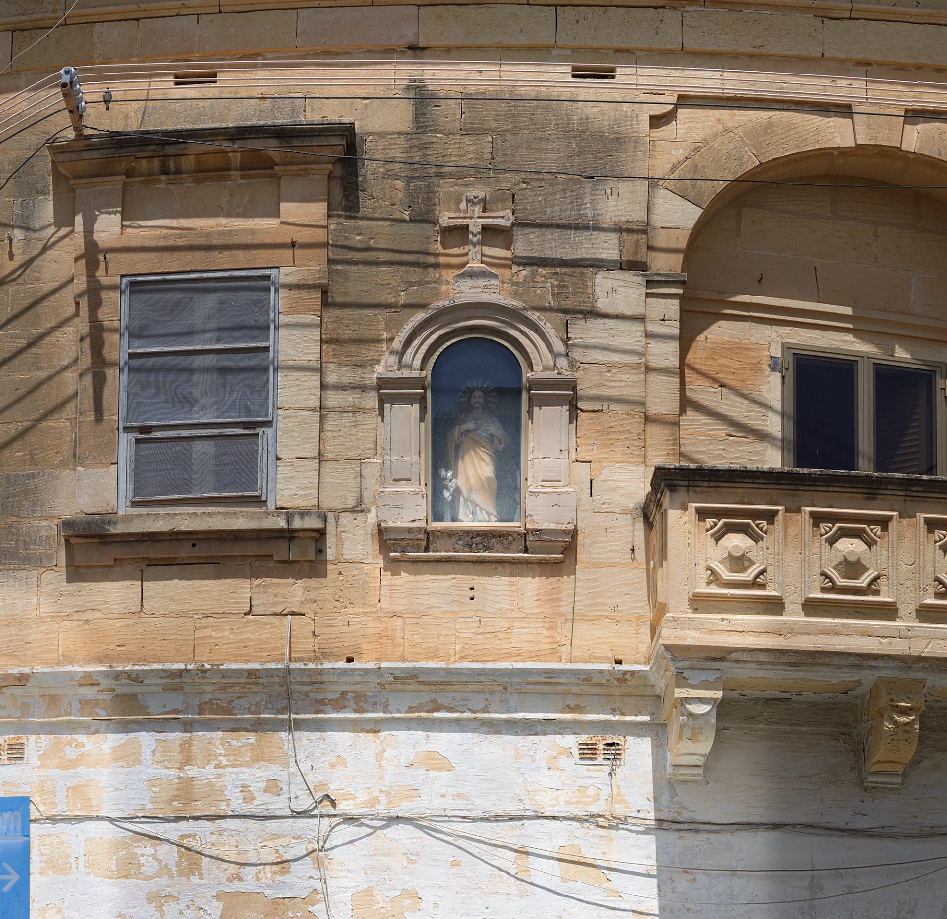View of the Niche of the Sacred Heart of Jesus on a weathered stone building facade, situated between a window and a stone balcony.