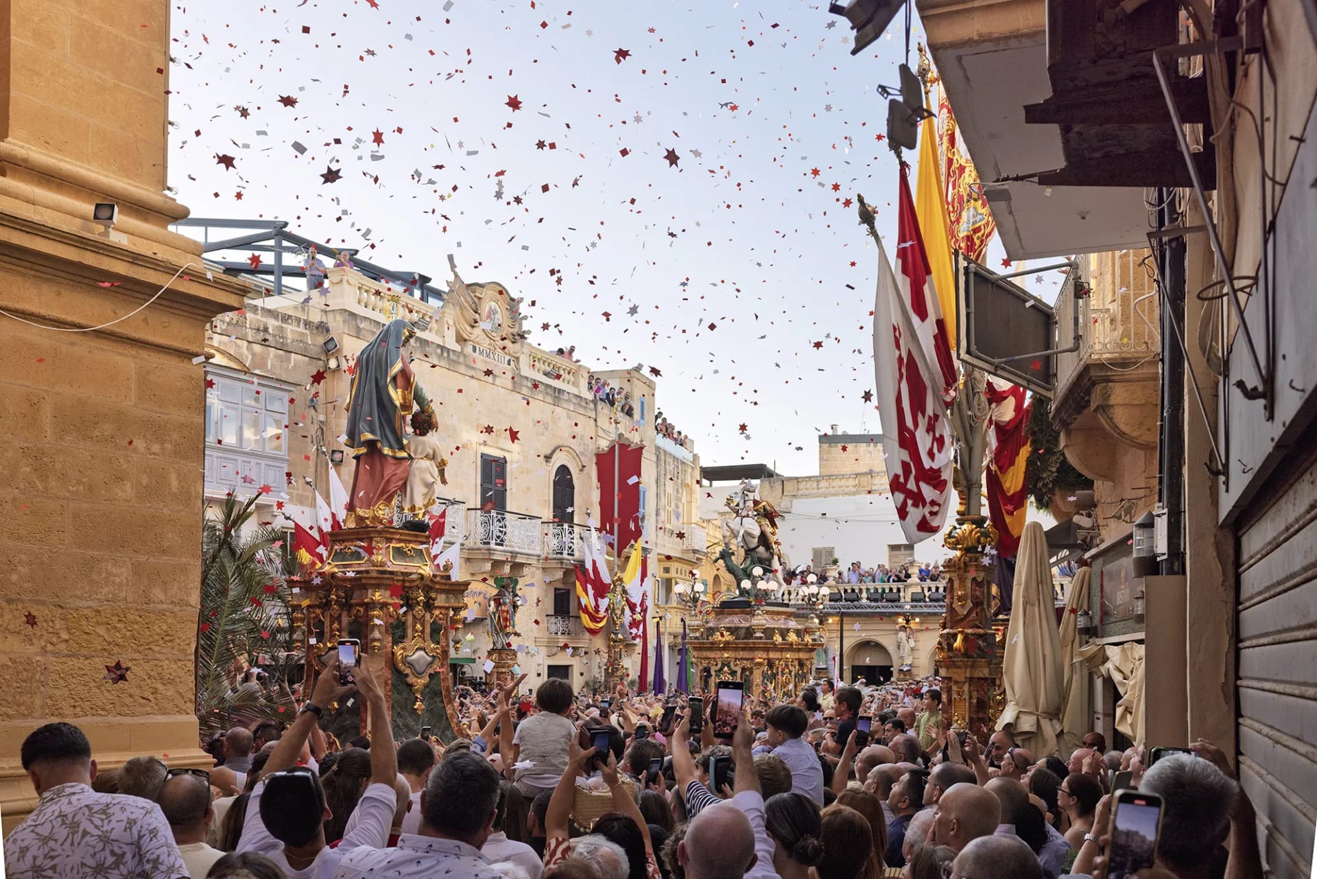 Crowds gather in Victoria, Gozo during the Festival of St George as statues of saints are paraded through confetti-filled streets, with flags, balconies, and onlookers adding to the vibrant celebration atmosphere.