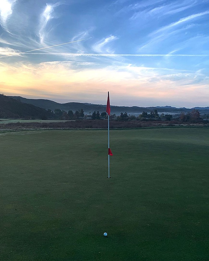 Beautiful sunset/sunrise view of a putting green with red flag, featuring dramatic sky with contrails and silhouetted mountains in the background.