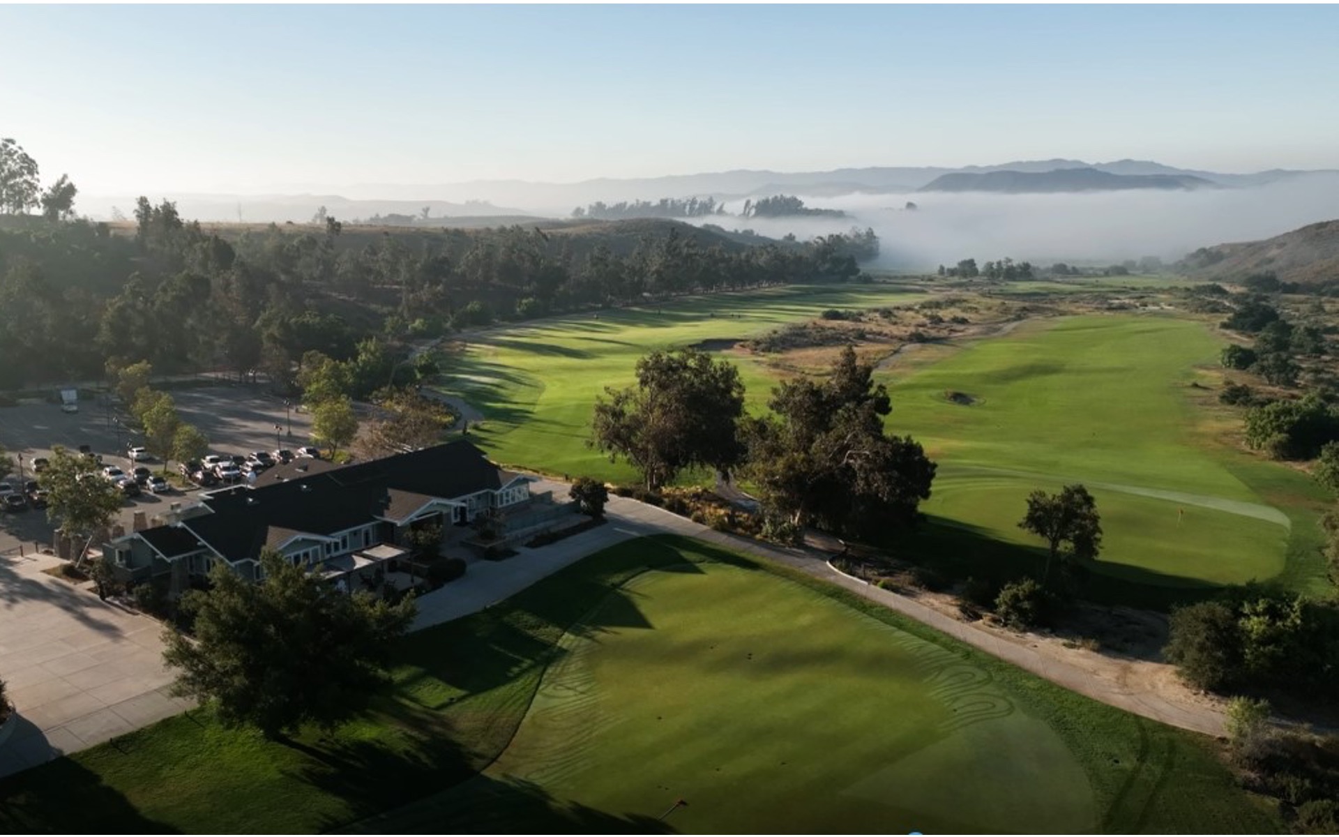 Early morning aerial view of the clubhouse facility with parking area, surrounded by golf course fairways and misty valley beyond.