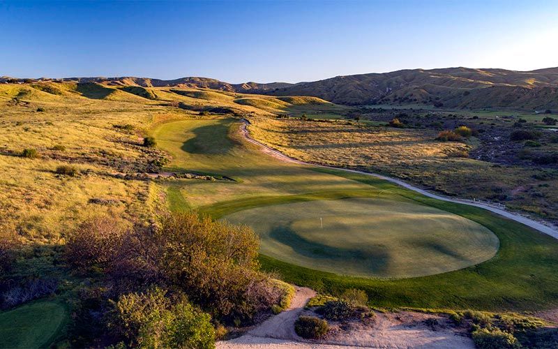 Ground-level view of a putting green surrounded by golden desert hills and natural vegetation in warm afternoon light.