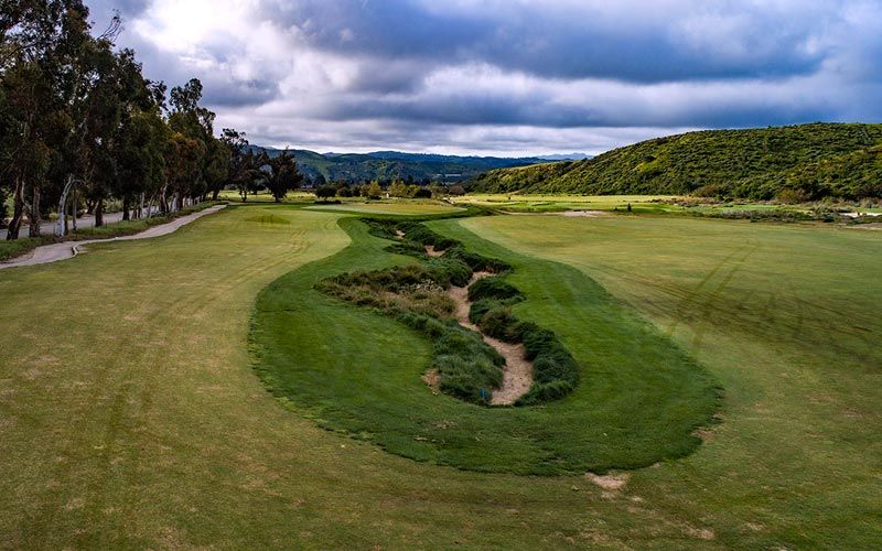 Wide fairway view with distinctive S-curved sand bunker cutting through the green under dramatic cloudy skies.